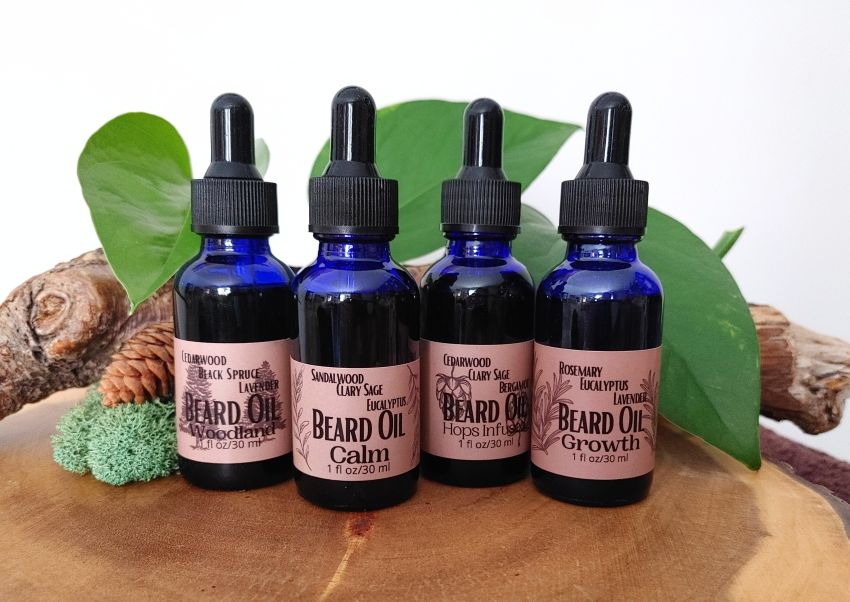 Four bottles of beard oil with droppers on a wooden surface with green leaves and natural elements.