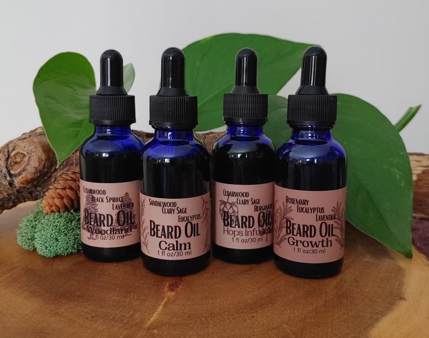 Four bottles of beard oil with labels on a wooden surface with green leaves in the background.