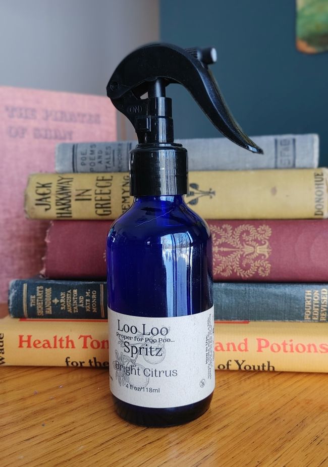 Blue spray bottle labeled 'Loo Loo Spritz' on a wooden surface with books in the background.