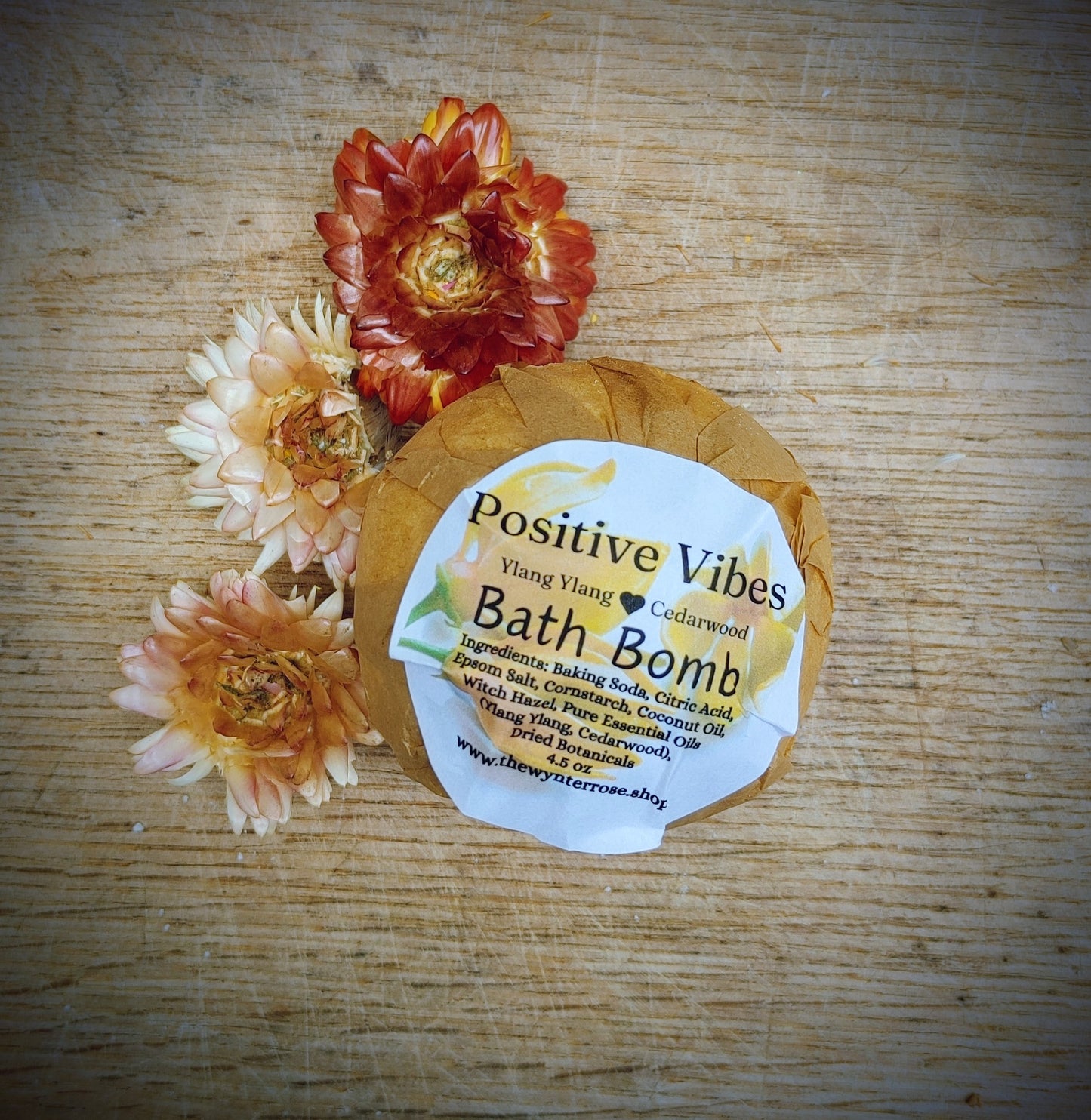 Bath bomb with a label reading 'Positive Vibes' surrounded by flowers on a wooden surface