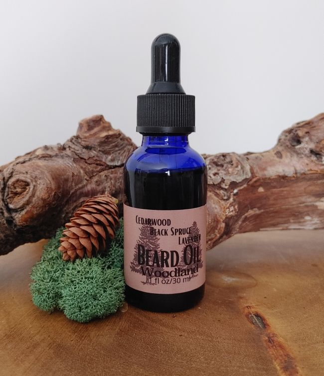 Bottle of 'The Woodland' beard oil with a dropper on a wooden surface with a pine cone and green moss.