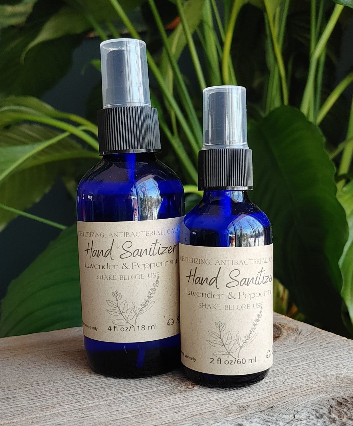 Two blue bottles of The Wynter Rose Lavender Peppermint Hand Sanitizer (4 oz and 2 oz), made with lavender and peppermint essential oils, rest on a wooden surface with green plants in the background.