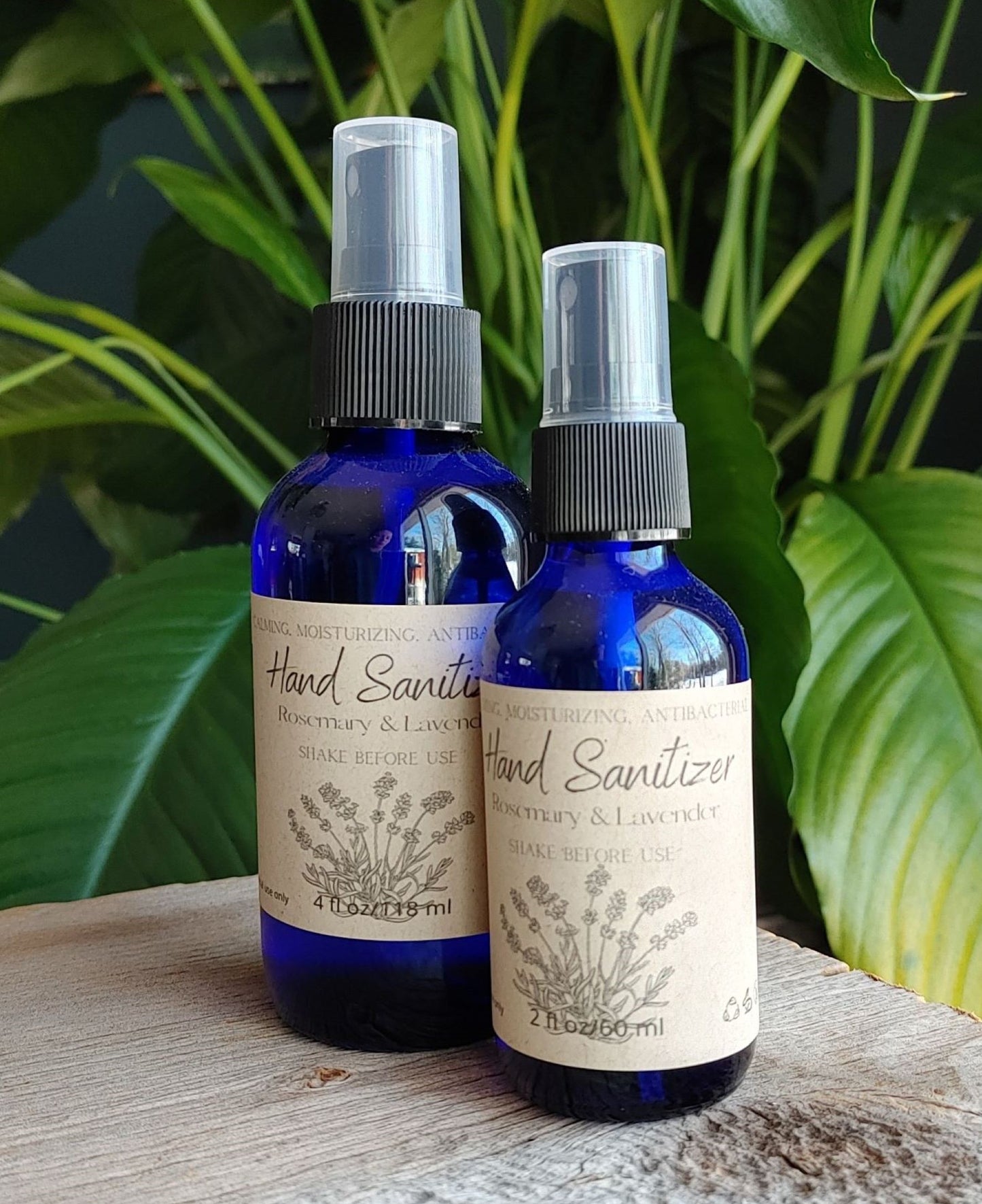 Two blue bottles of The Wynter Rose Lavender Rosemary Hand Sanitizer Spray with black tops and beige labels featuring botanical illustrations sit on a wooden surface, framed by green leafy plants in the background.