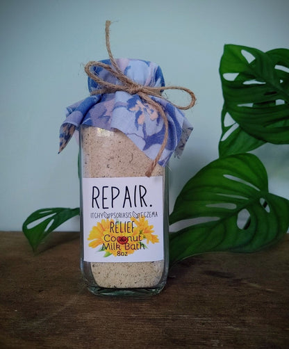 repair body soak for eczema relief Bottle of coconut milk bath with a decorative label on a wooden surface with green leaves in the background.