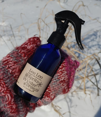Blue spray bottle labeled 'Loo Loo Spritz Wynter Breeze' on a textured surface with a red knitted item.