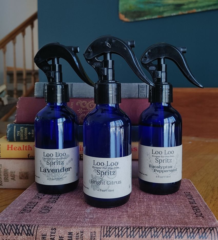 Three blue spray bottles labeled 'Loo Loo Spritz' on a textured surface with books in the background.