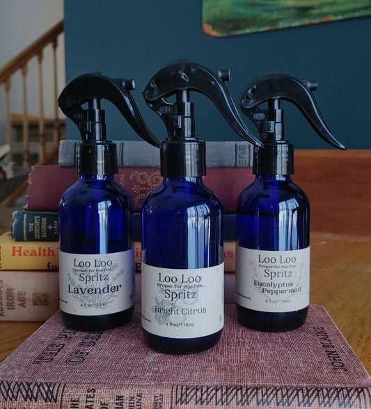Three blue spray bottles labeled 'Loo Loo Spritz' on a textured surface with books in the background.
