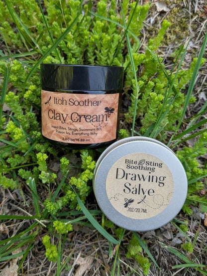 Two containers of natural remedies, 'Itch Soother Clay Cream' and 'Drawing Salve', on grass.