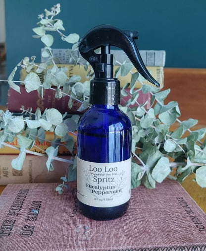 Blue spray bottle labeled 'Loo Loo Spritz' with eucalyptus and peppermint on a surface with greenery.