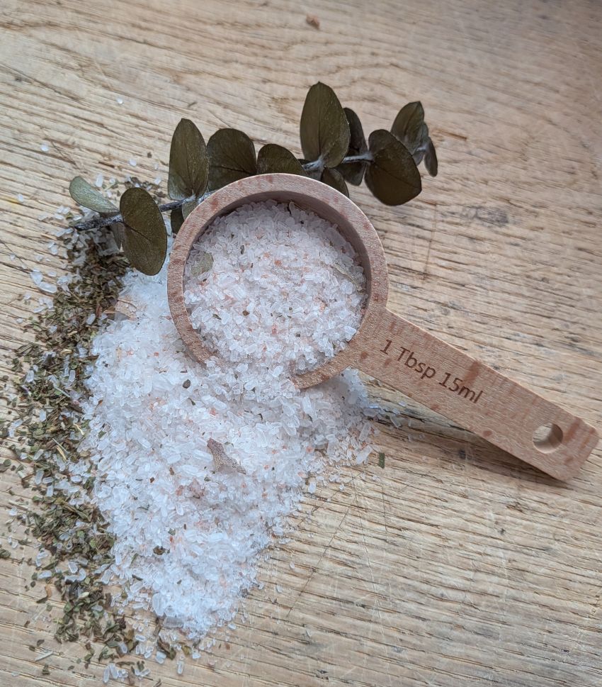 A wooden measuring scoop filled with coarse white and pink Himalayan salt, with loose salt spilled beside it and dried eucalyptus leaves on a light wood surface. The scoop handle is engraved with '1 Tbsp 15ml'.