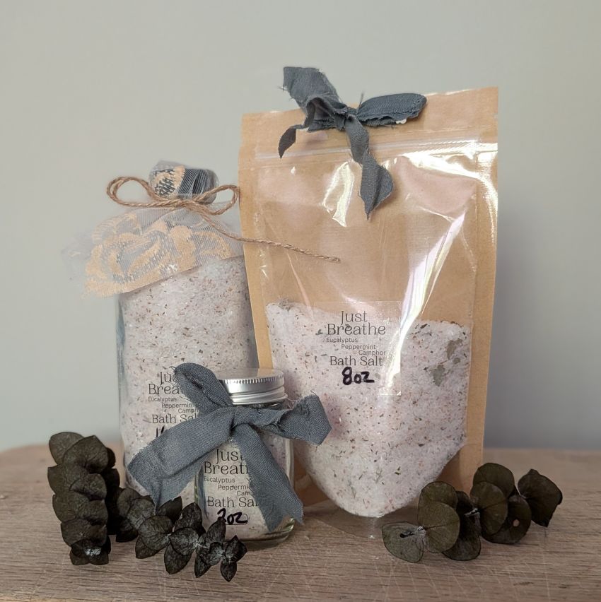 Two bags of bath salts displayed on a wooden surface against a neutral gray background. One bag is kraft paper with a gray ribbon, and the other is a clear jar with a silver lid and a gray ribbon.