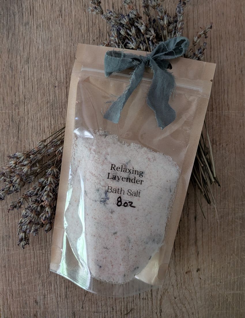 A 8 oz bag of Relaxing Lavender Bath Salt in a clear stand-up pouch with a kraft paper backing and a dusty blue ribbon bow, displayed on a wooden surface with dried lavender sprigs.