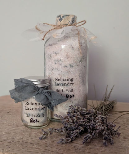 Two glass jars of lavender bath salt on a wooden surface with dried lavender sprigs. The larger jar is filled with pale pink and white bath salt and tied with a sheer white ribbon and twine.