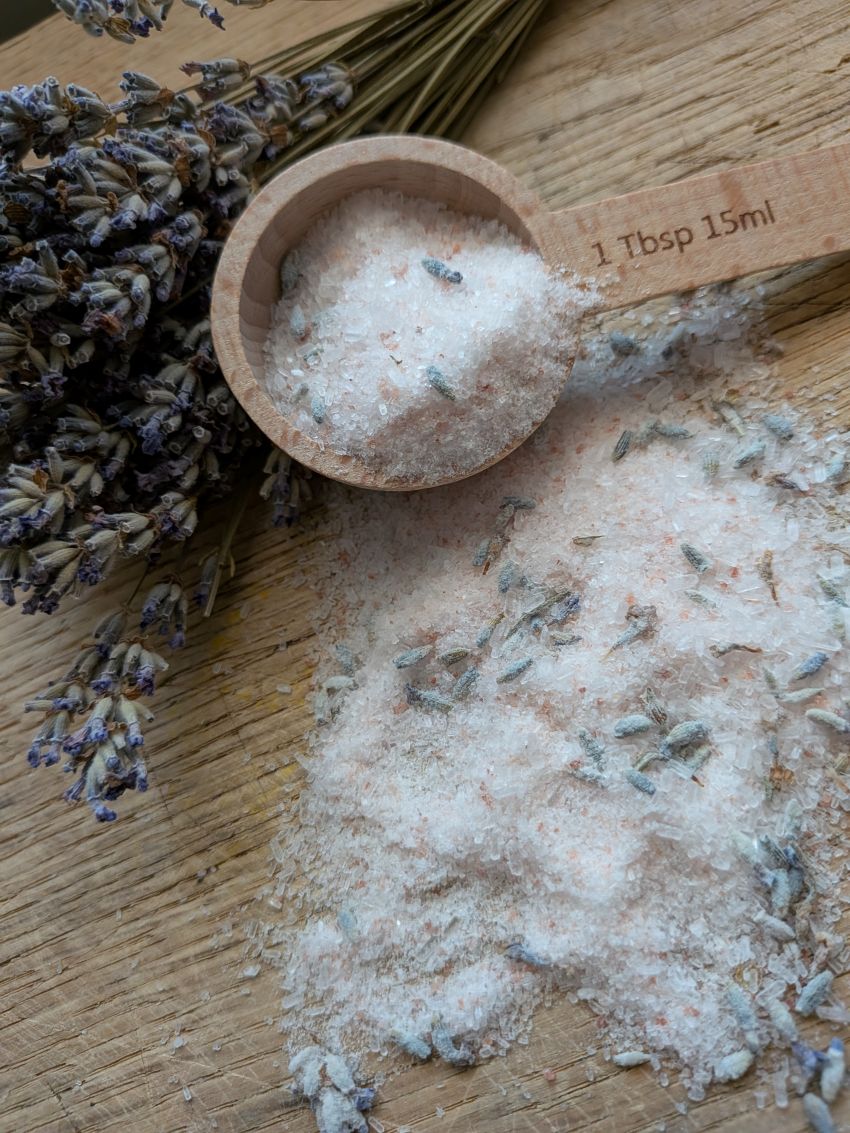 Lavender salt bath mixture in a wooden measuring scoop with '1 Tbsp 15ml' printed on it, spilled onto a wooden surface next to dried lavender sprigs.