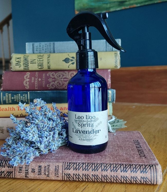 Blue spray bottle labeled 'Loo Loo Spritz Lavender' on a stack of books with lavender flowers.