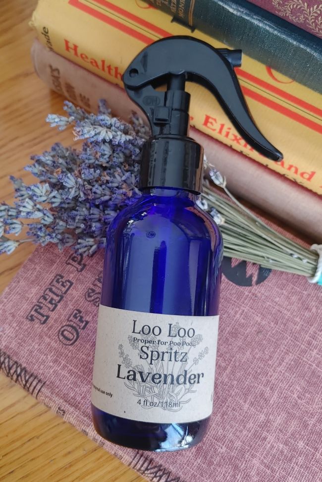 Blue spray bottle labeled 'Loo Loo Spritz Lavender' on a wooden surface with books in the background.