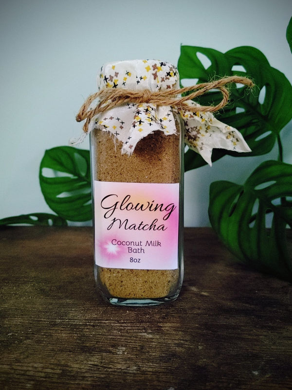 Jar of 'Glowing Matcha' coconut milk bath on a wooden surface with green leaves in the background