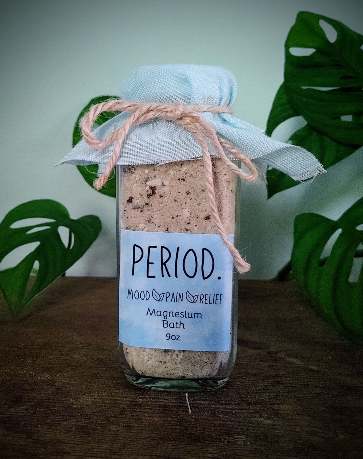 Bottle of 'Period' magnesium bath salt menstrual PMS symptom relief bath soak with a label on a wooden surface with green leaves in the background.