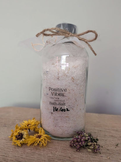 clear glass bottle with positive vibes therapeutic bath sale on a wooden board with dried florals around it. vegan body soak made by The Wynter Rose