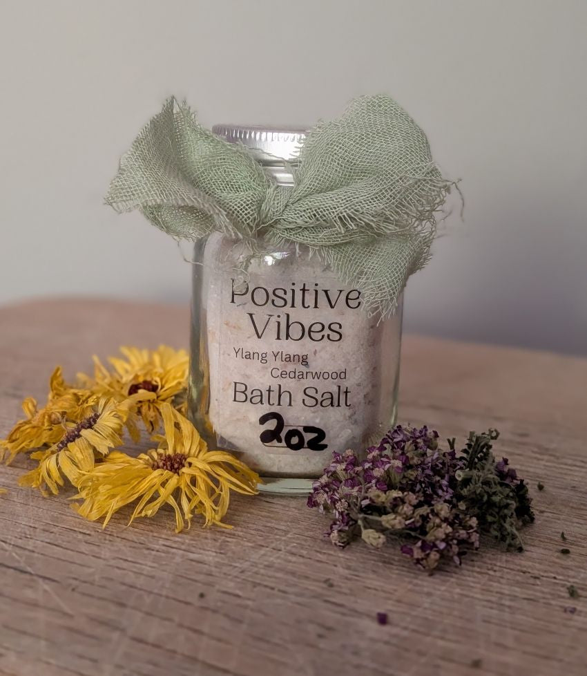 A glass jar of Positive Vibes Bath Salt sits on a rustic wooden surface, decorated with a sage green burlap bow. The jar is styled with dried yellow flowers and purple lavender sprigs, set against a soft, neutral background.