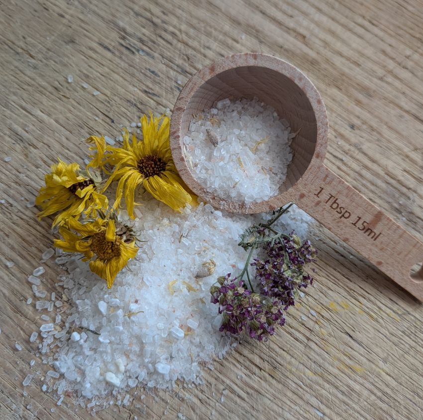 White bath salts mixed with dried yellow and purple flowers, displayed on a wooden surface next to a wooden measuring scoop.