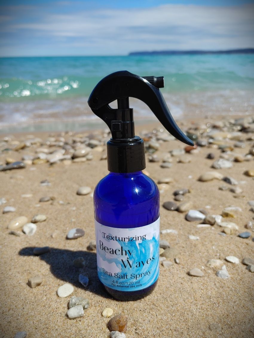 sea salt beachy waves hair spray for beach vibe mermaid hair housed in a blue glass bottle with black trigger spray top sandy michigan beach sleeping bear dunes backdrop