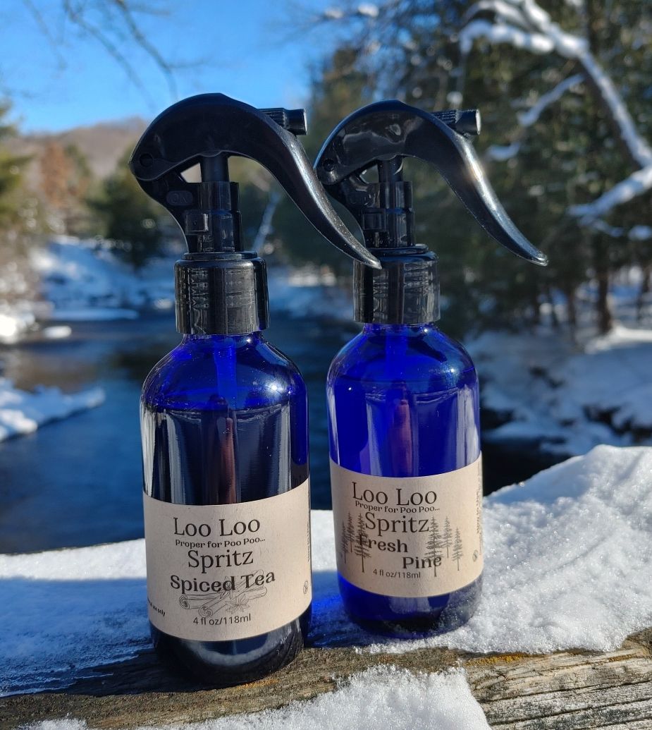 Two blue spray bottles labeled 'Loo Loo Spritz' on a snowy surface with a winter landscape in the background.