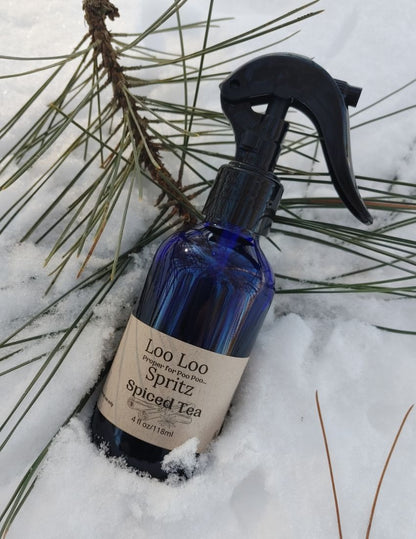 Blue spray bottle labeled 'Loo Loo Spritz Spiced Tea' in the snow with pine branches.