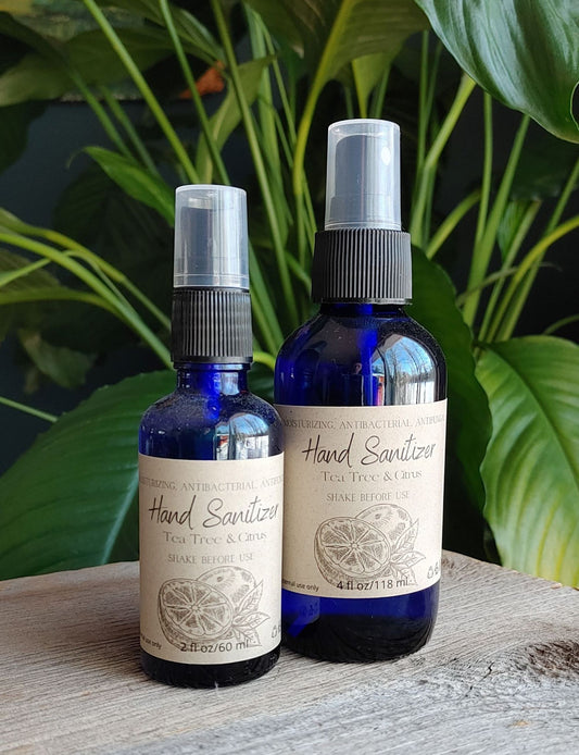 Two blue bottles of The Wynter Rose Tea Tree Citrus Hand Sanitizer, featuring beige labels and enriched with tea tree oil, are shown in front of green plants. The bottles come in two sizes.